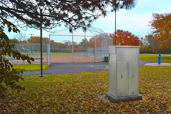 A silver commercial meter pedestal next to a baseball diamond taking up less space than the strut and backboard.