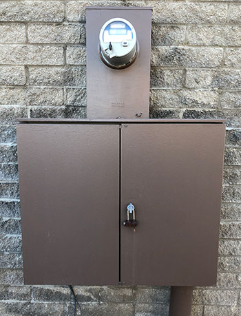 A brown meter socket is mounted top of a brown CT cabinet against a light brick wall.